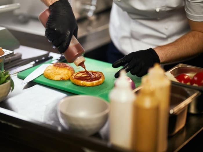 Food truck événementiel Nantes - Traiteur Événementiel Nantes & Vitré | Restonomade Chef en gants noirs préparant un burger dans un food truck, ajoutant de la sauce sur un pain brioché posé sur une planche verte.